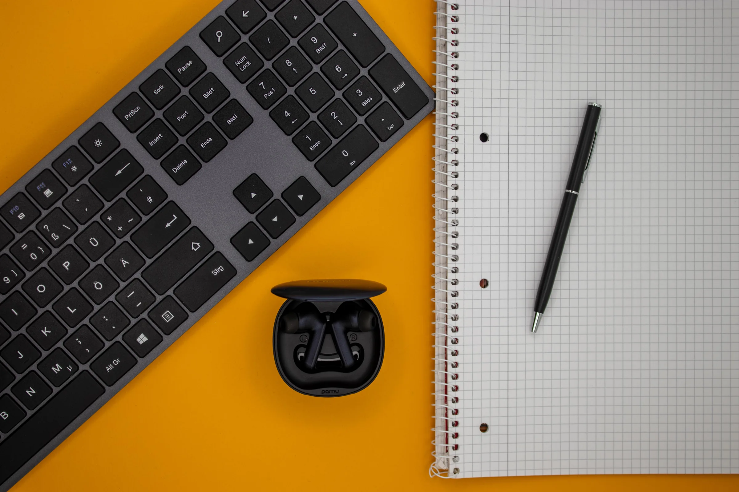 Flatlay auf orangem Grund: Tastatur, Ladecase mit Ohrhörern, Notizblock und Kugelschreiber
