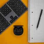 Draufsicht Auf Eine Computer-Tastatur, Ein Notizbuch Mit Kugelschreiber Und Kabellose Ohrhörer Auf Einem Gelben Hintergrund.